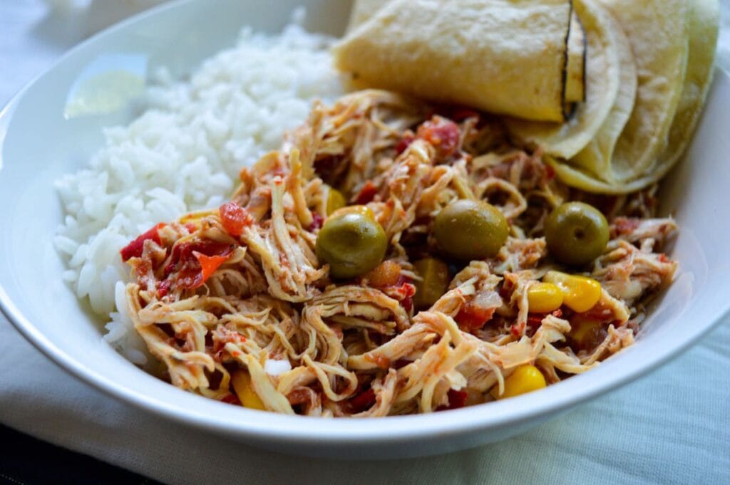 Shredded chicken covered in a rich tomato sauce with olives is served with white rice and tortillas in a white bowl.