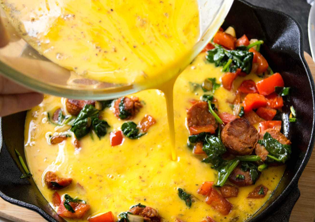 Egg mixture being poured over a skillet with veggies in it.