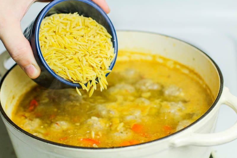 Uncooked orzo being poured into saffron albondigas soup