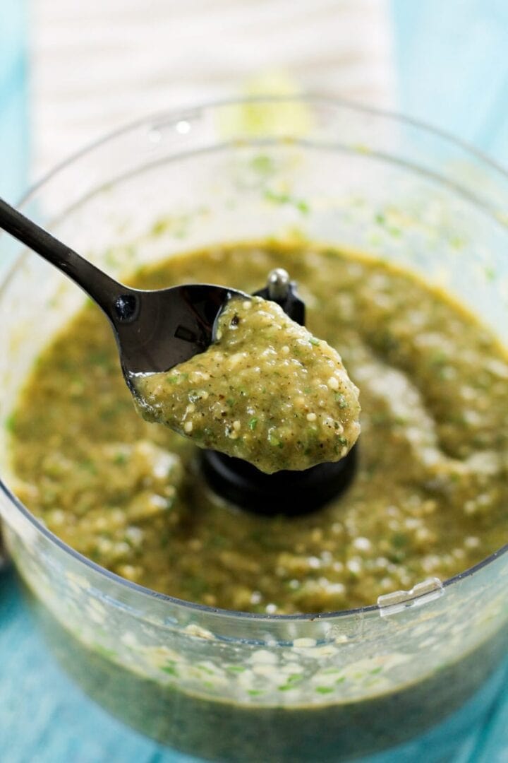 Freshly made salsa verde is in a food processor with a blue background and a black spoon holding up a small amount to show viewers.