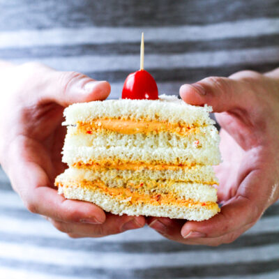 man holding 3 sliced sandwiches with a tomato in a toothpick