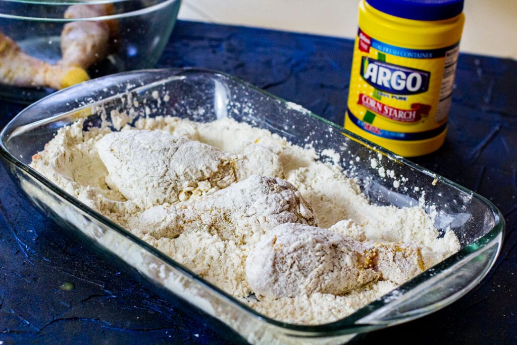 Raw coated chicken in a large glass container filled with flour.