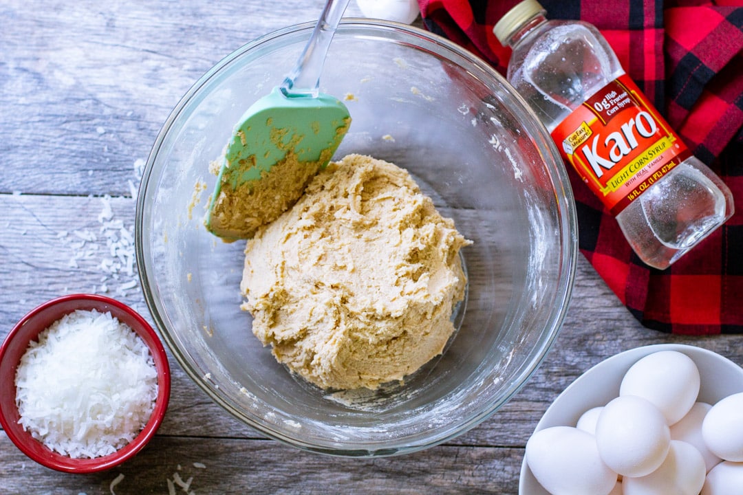 Raw coconut cookie dough mixture in a glass bowl with a green rubber spatula.