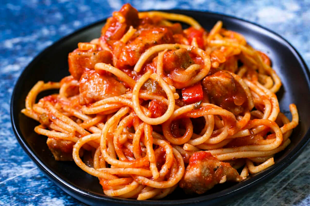 Close of shop of a black plate filled with cooked spaghetti with tomato sauce and chunks of pork and veggies.