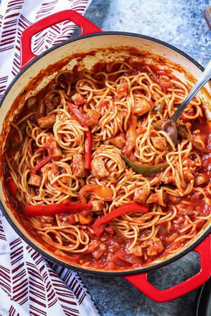 A red pot is full of spaghetti with cut pork chops and bell peppers on a blue background.