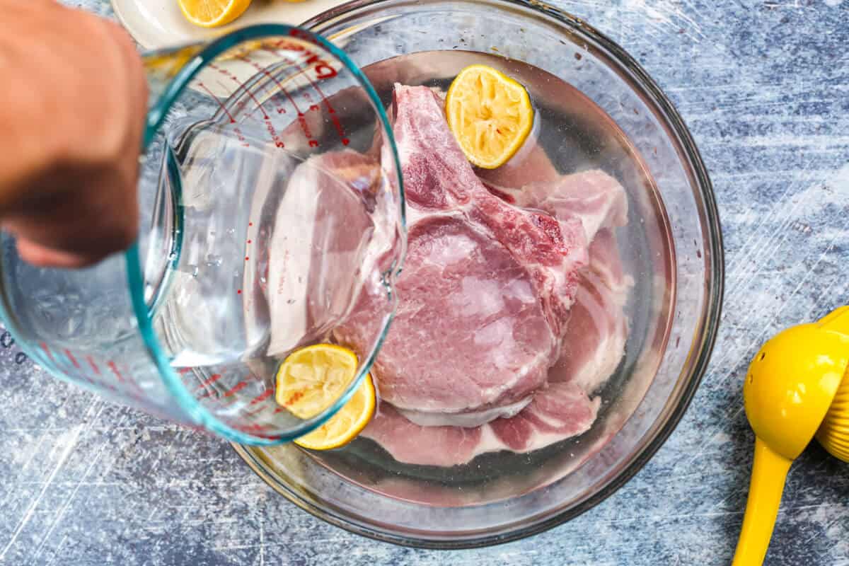 Large bowl filled with raw pork chops brining with water and lemons.