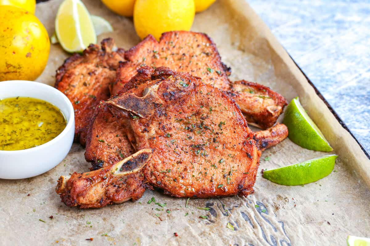 Cooked pork chops on parchment paper, garnished with parsley flakes and surrounded by wedges of lime.