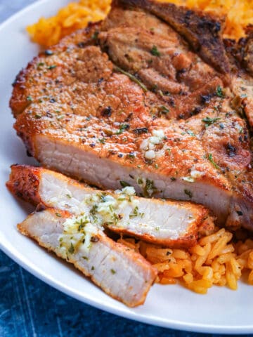 A crispy fried pork chop that is sliced is laying over a bed of Puerto Rican rice on a white plate.