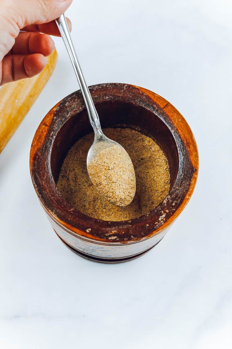 A mortar and pestle filled with seasoning blend with a metal spoon holding some of the spices.