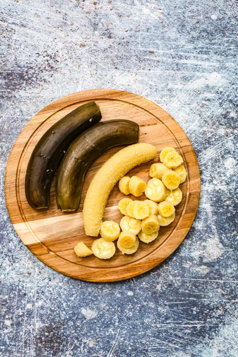 A wooden cutting board, showing the stages of prepping cooked green bananas for escabeche.