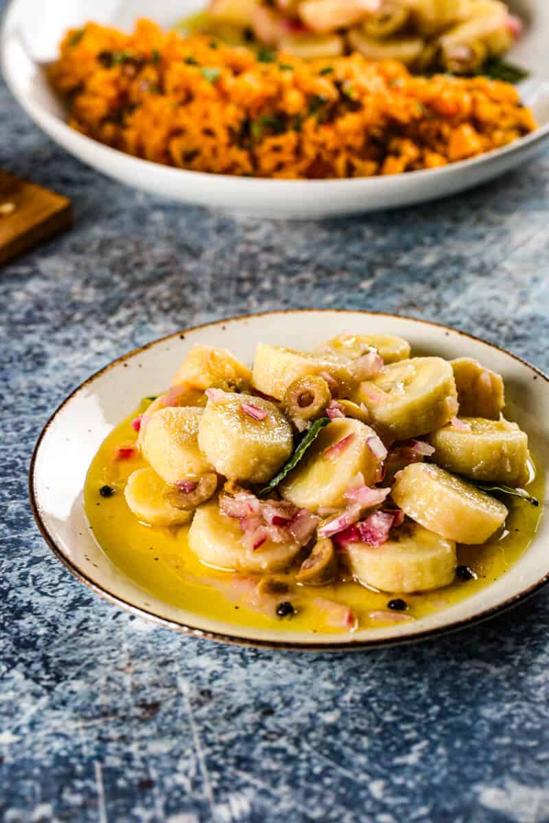 Escabeche con guineos in a zesty pickled onion sauce with a plate of Spanish rice in the background.