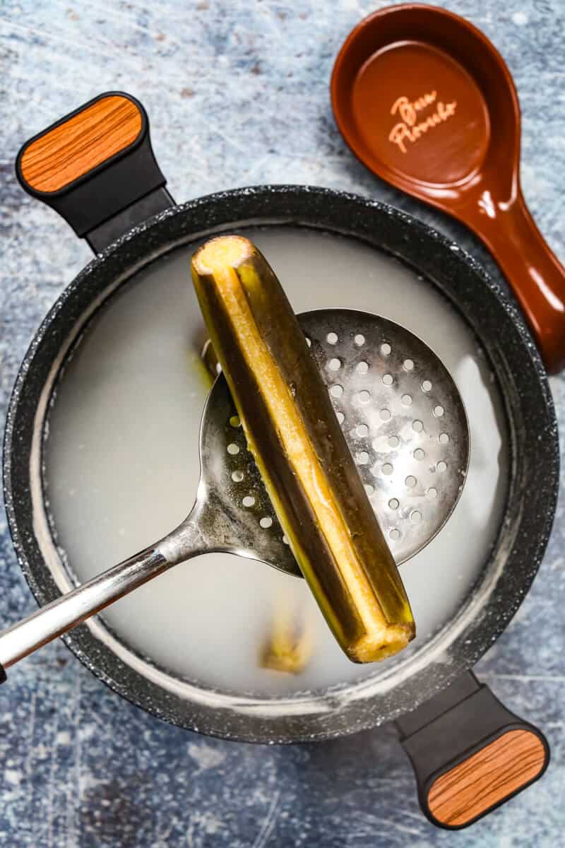 A metal splotted cooking spoon is holding a cooked green banana with a pot of milky water below it.