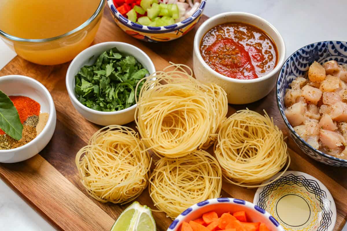 Ingredients for sopa de fideo are on a wooden cutting board in separate small bowls.