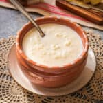 A bowl filled with warm and creamy rice milk soup that's known as arroz con leche is centered over an artisan style small bowl with a spoon sitting inside of the soup.