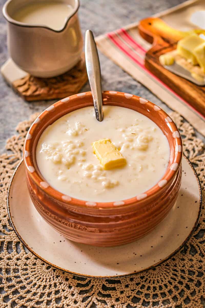 A small brown bowl is filled with creamy white rice milk soup that has a spoon sunk into it and a dollop of butter over it.
