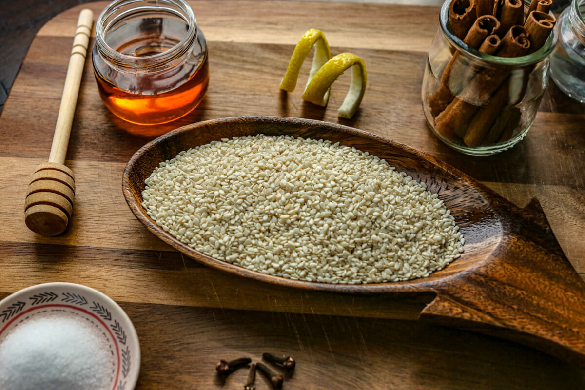 White sesame seeds are in a brown bowl with cinnamon sticks, honey, and a lemon rind near it on a wooden cutting board.