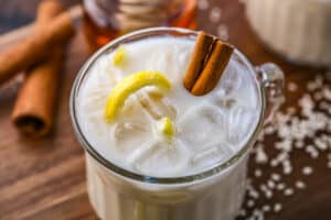 A small glass mug is filled with Puerto Rican horchata with a cinnamon stick and lemon rind on top of a wooden board.