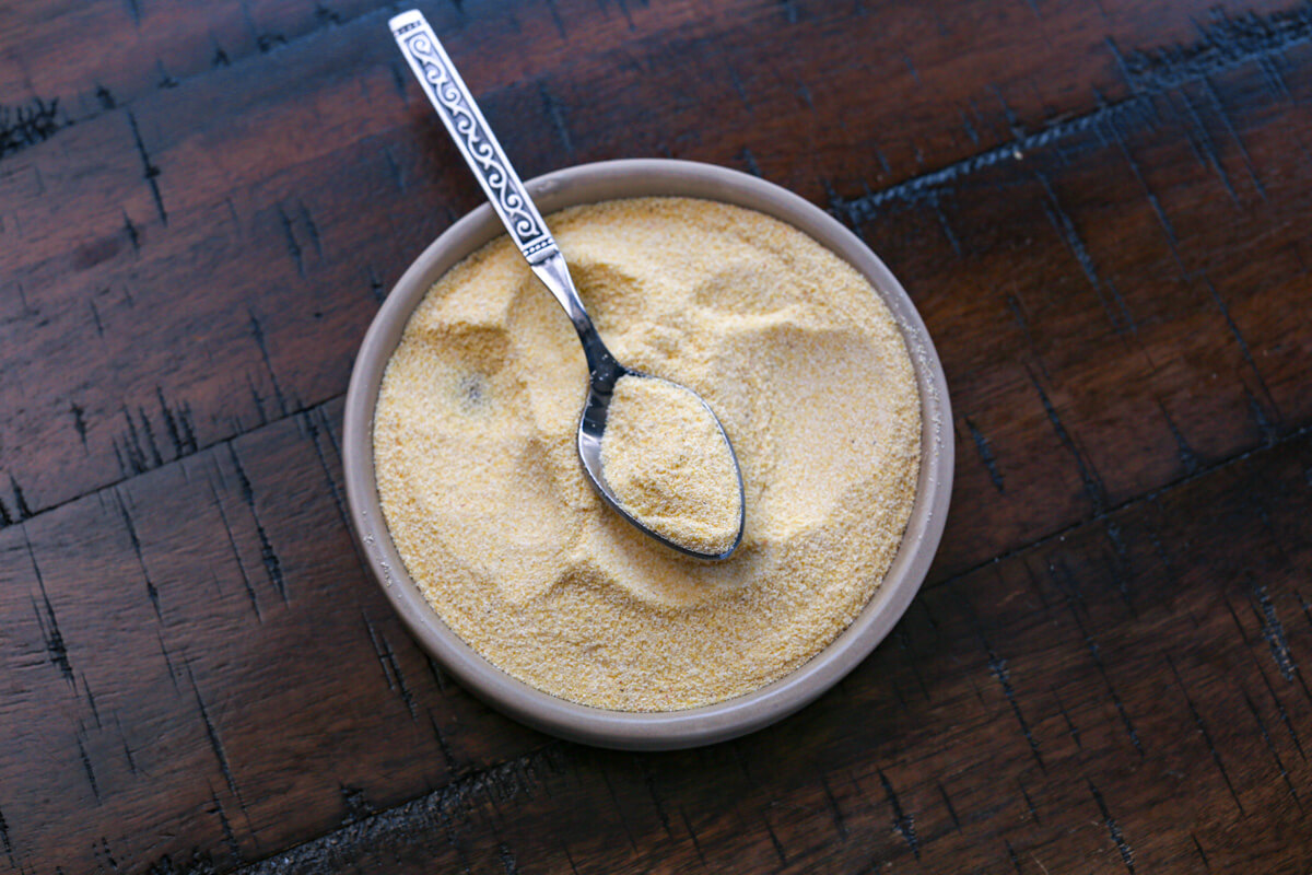 A small ceramic bowl is filled with dry cornmeal flour with a metal spoon in the center. The background is wooden.