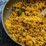 Overhead shot of an aluminum pan full of yellow rice with pigeon peas with a bay leaf and a wooden spoon.