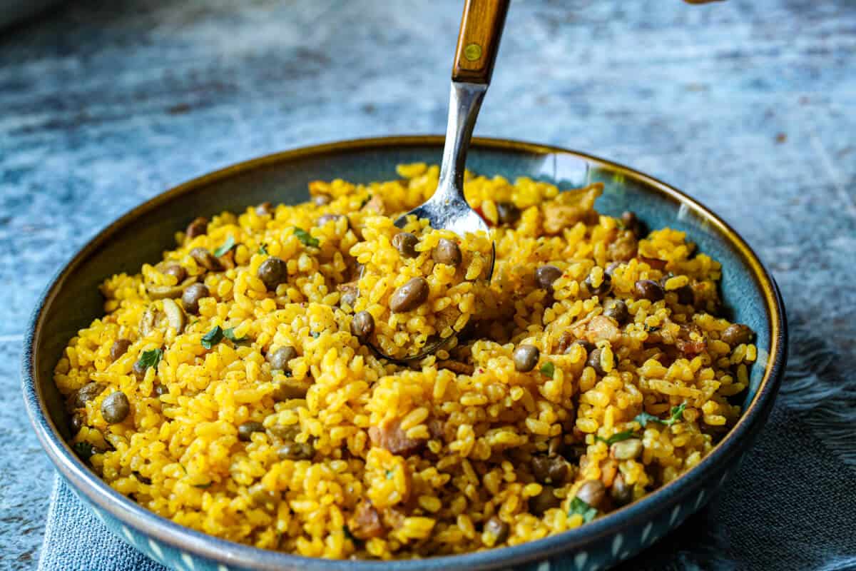 A blue bowl is filled with arroz con gandules and there is a wooden handled spoon holding up some of the yellow rice with pigeon peas with a blue background.