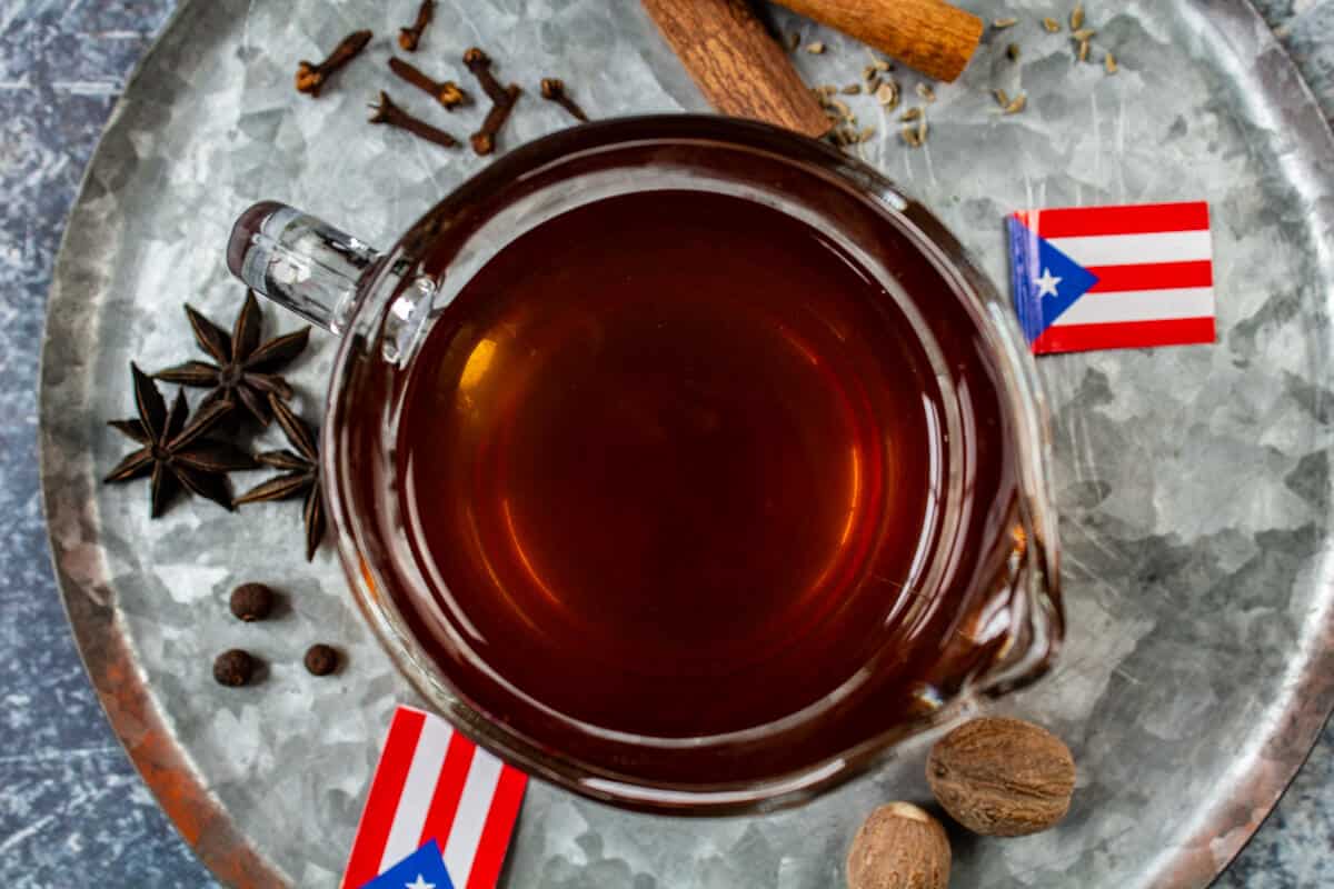 An overhead shot of an amber colored spiced tea for coquito in a glass cup on a metal plate with various spices, nutmeg, and small Puerto Rican flags.