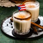 Close up shot of coquito in a glass cup with cinnamon on the rim and a Puerto Rican flag.