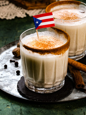 Close up shot of coquito in a glass cup with cinnamon on the rim and a Puerto Rican flag.