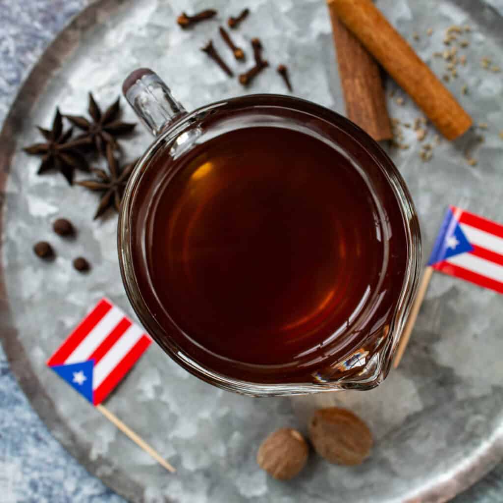 Overhead shot of spiced tea for coquito in a glass cup with Puerto Rico's flags, cinnamon sticks, cloves, anise, and spices in the background.