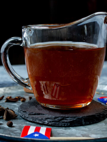 Tea for coquito is inside of a glass cup with a gray and blue background, spices, and Puerto Rican flags.