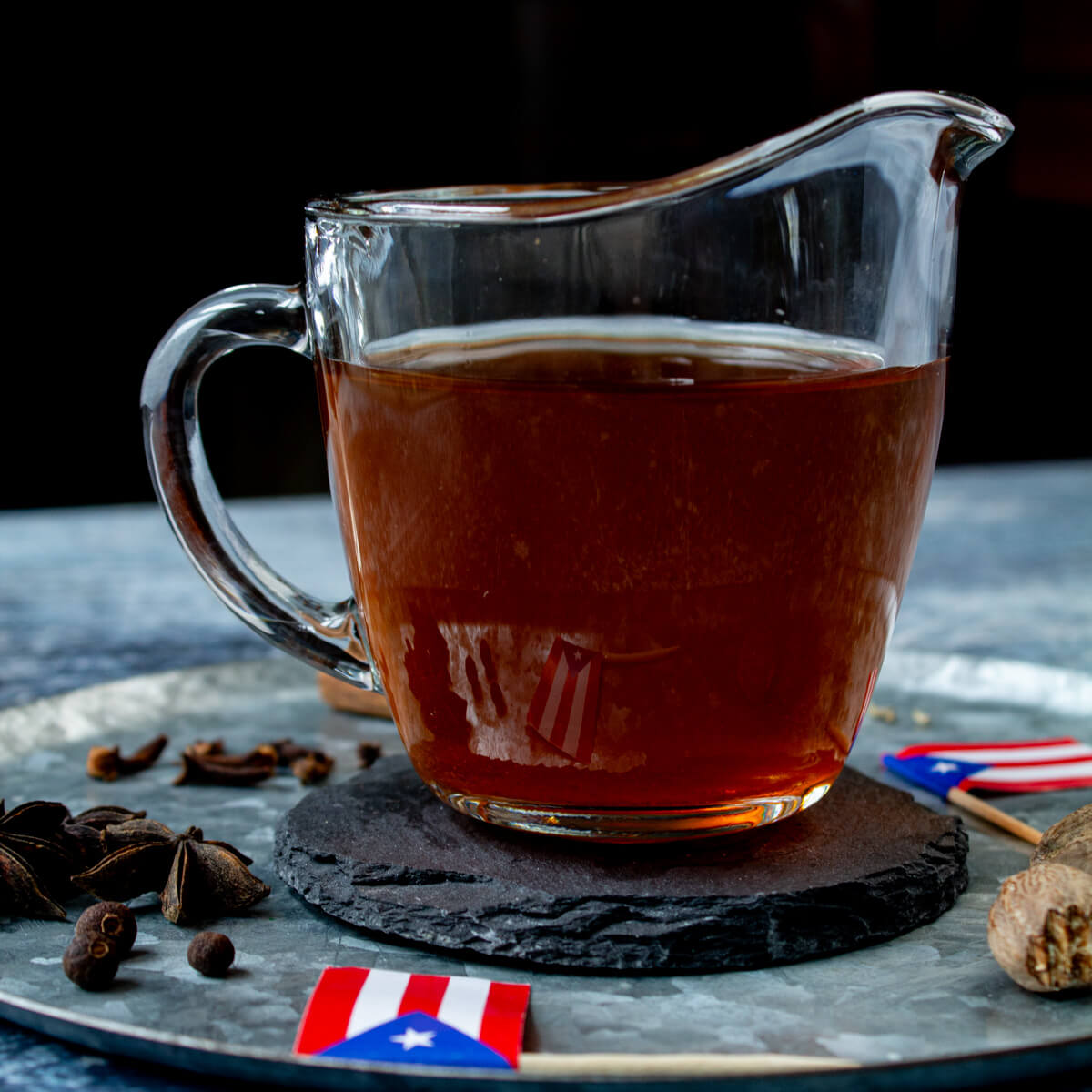 Tea for coquito is inside of a glass cup with a gray and blue background, spices, and Puerto Rican flags.