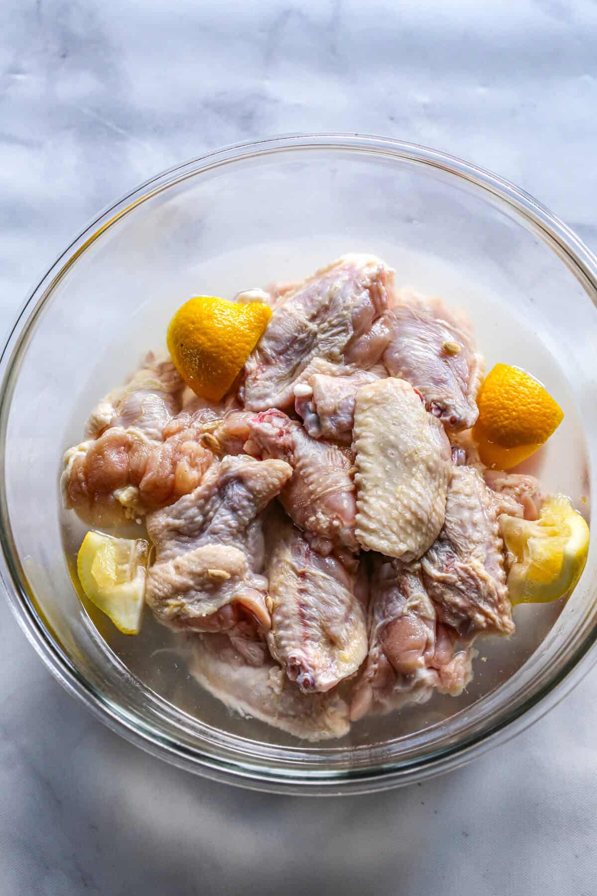 A glass bowl full of chicken wings brining in water, vinegar, with chunks of yellow lemons.