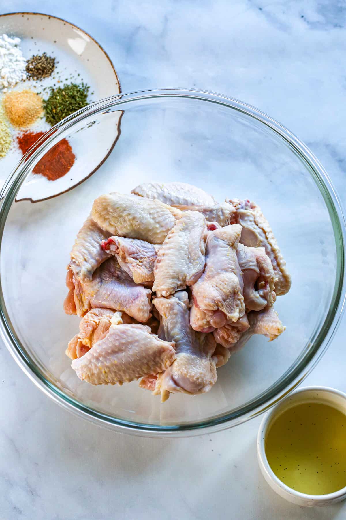 A glass bowl of chicken wings, with a small side bowl full of olive oil, and a plate on the top left hand side full of seasonings.