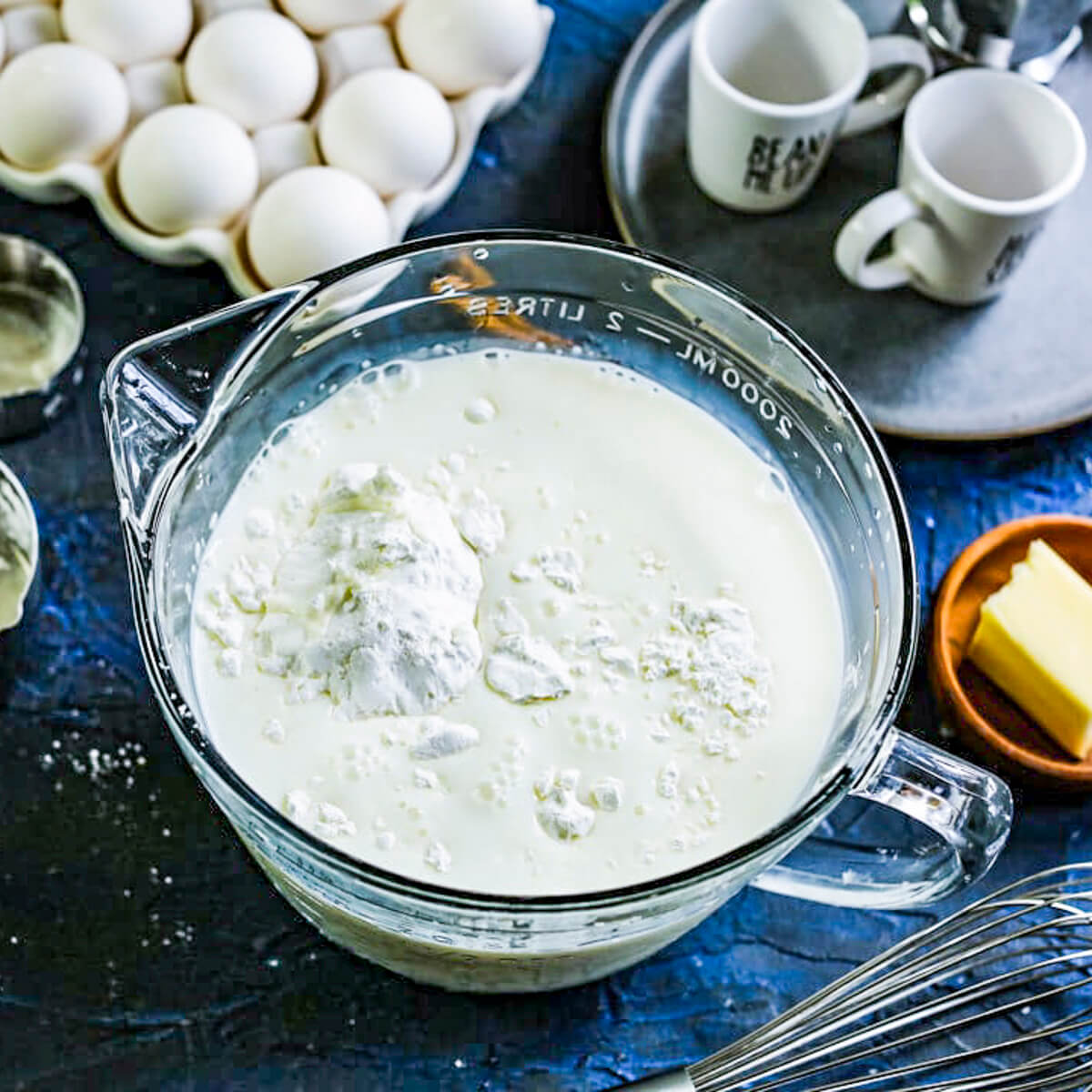 A glass mixing bowl is filled with milk and ingredients, with a blue background.