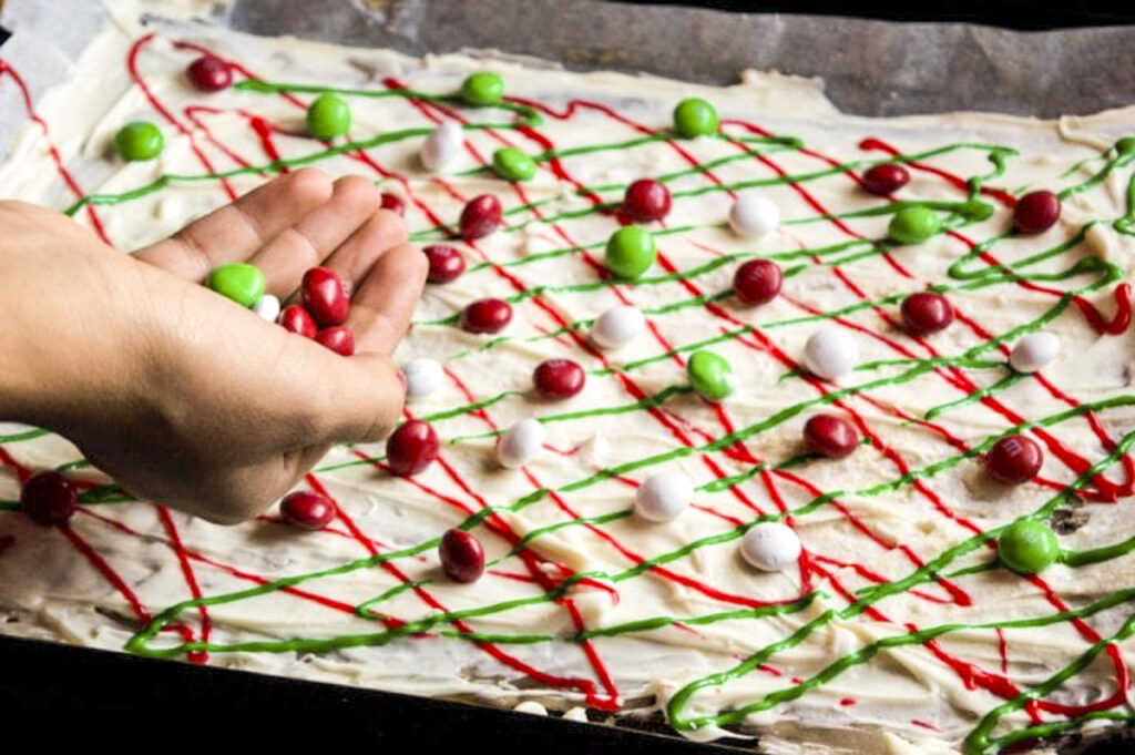 Child's hand is pouring white chocolate chips over the candy bark that has red and green swirl designs on it.
