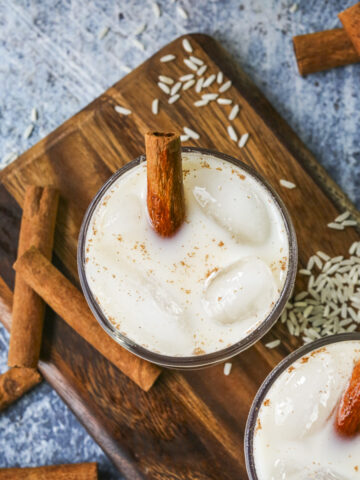 OVerhead shot of horchata in a glass cup with cinnamon sticks, ice, and grains of rice in the blue background.