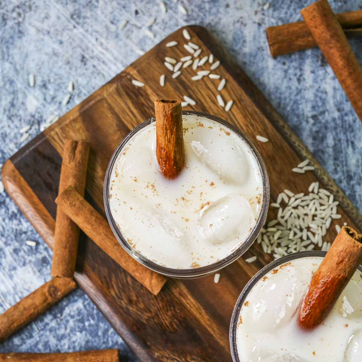OVerhead shot of horchata in a glass cup with cinnamon sticks, ice, and grains of rice in the blue background.