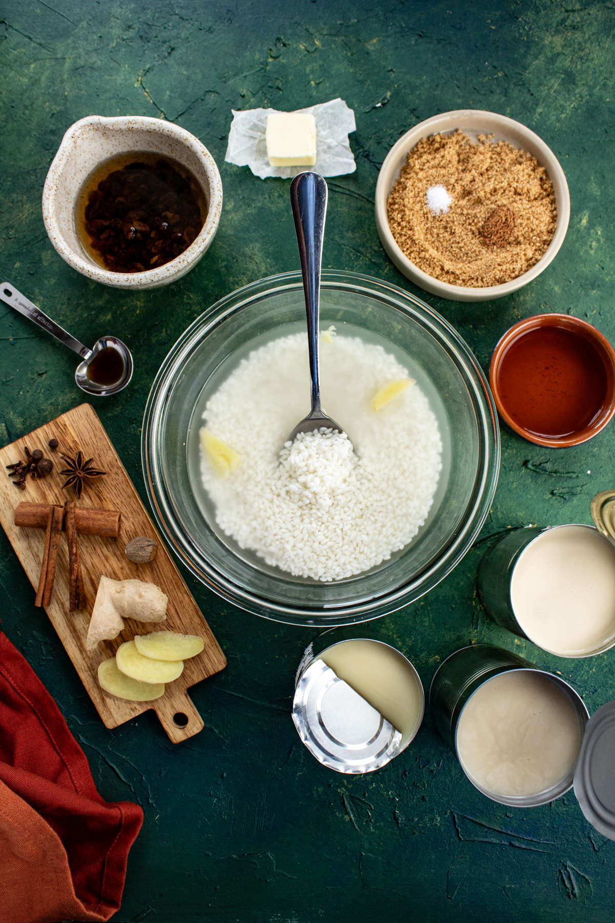 A green background is covered in an array of small dishes that hold ingredients for this arroz con dulce recipe, like rice, cinnamon sticks, etc.