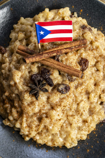 Golden rice pudding is topped with cinnamon sticks and a Puerto Rican flag, on a dark gray plate.
