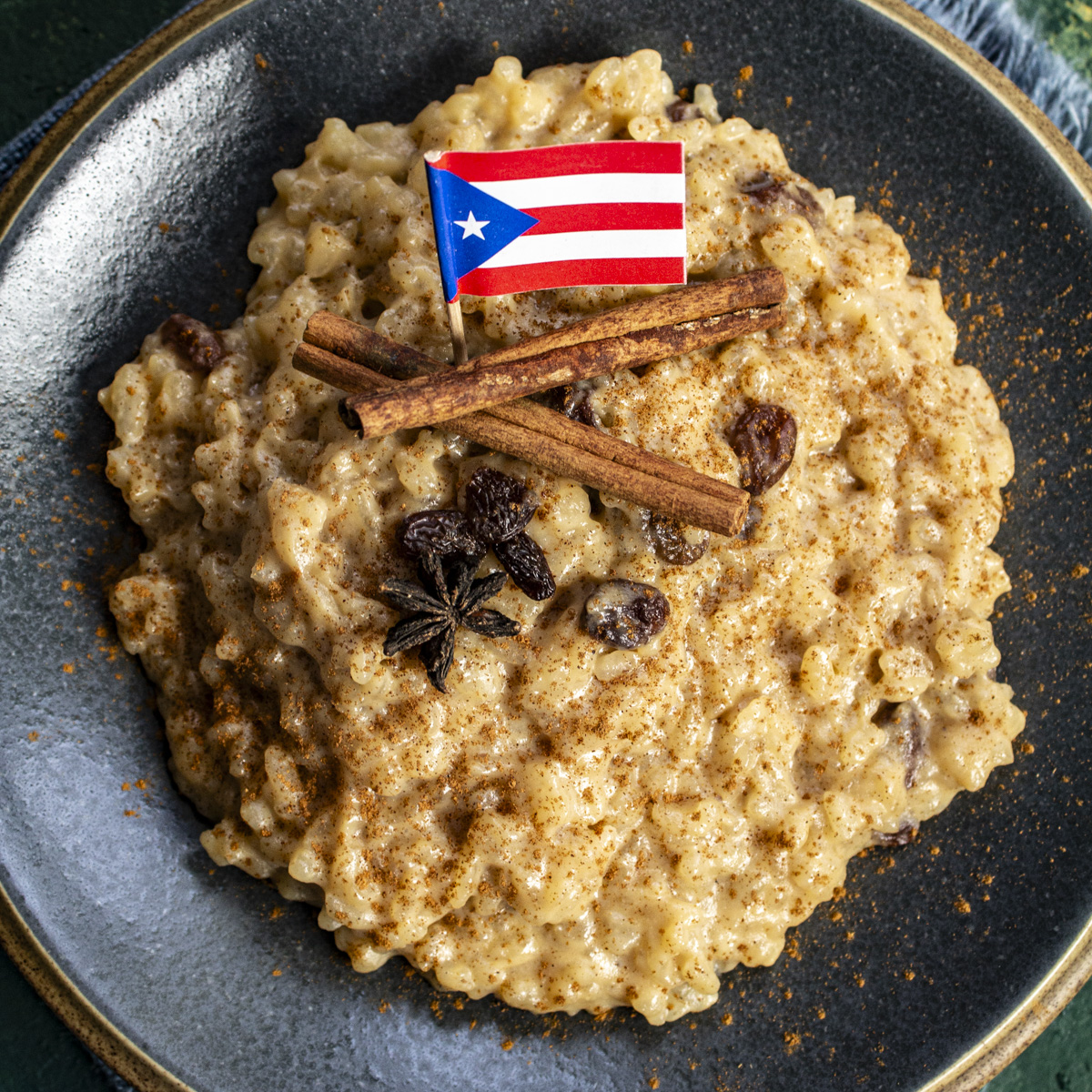 Golden rice pudding is topped with cinnamon sticks and a Puerto Rican flag, on a dark gray plate.