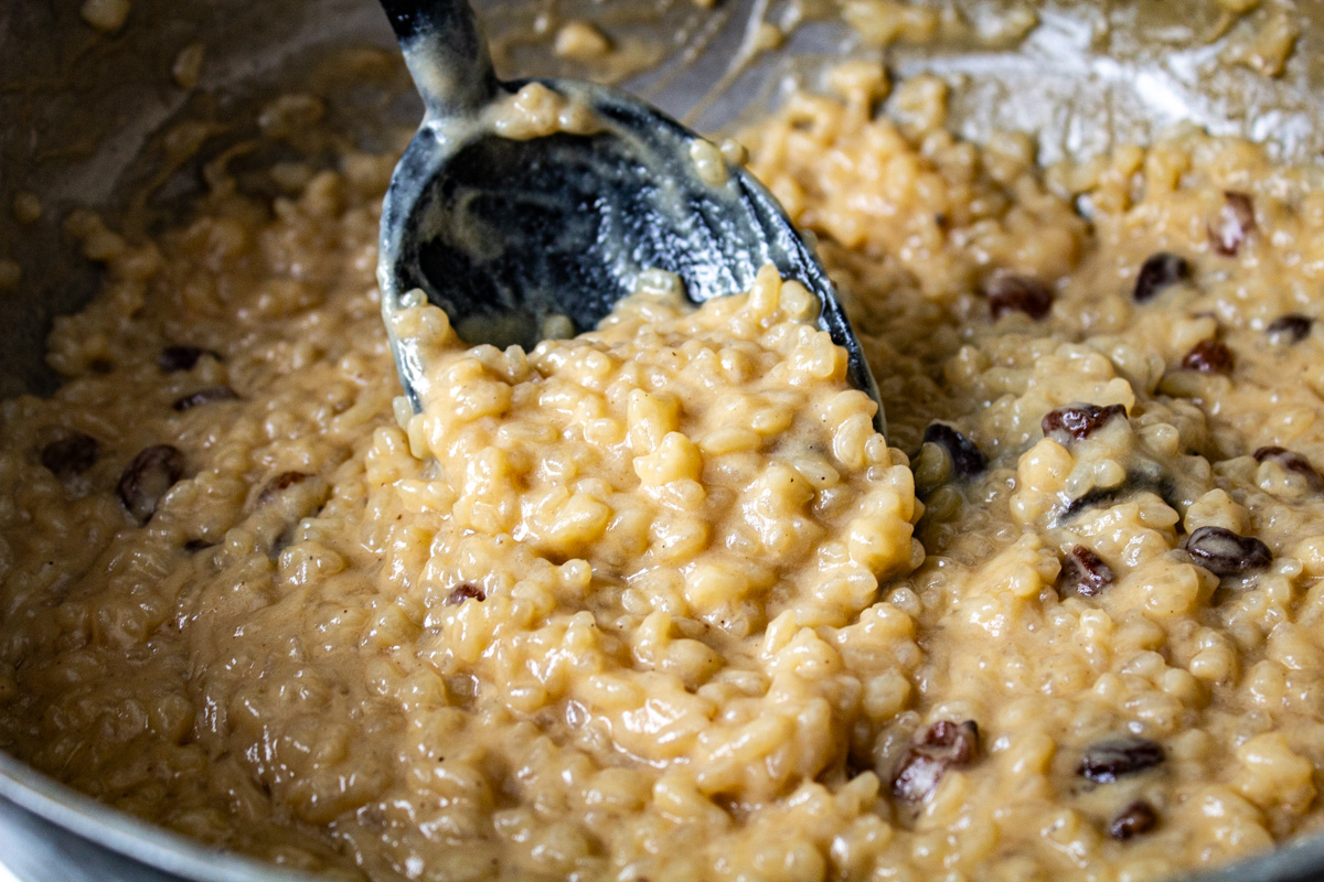 Close up of a black spoon holding up finished, thick and creamy rice pudding in a cast iron pot.