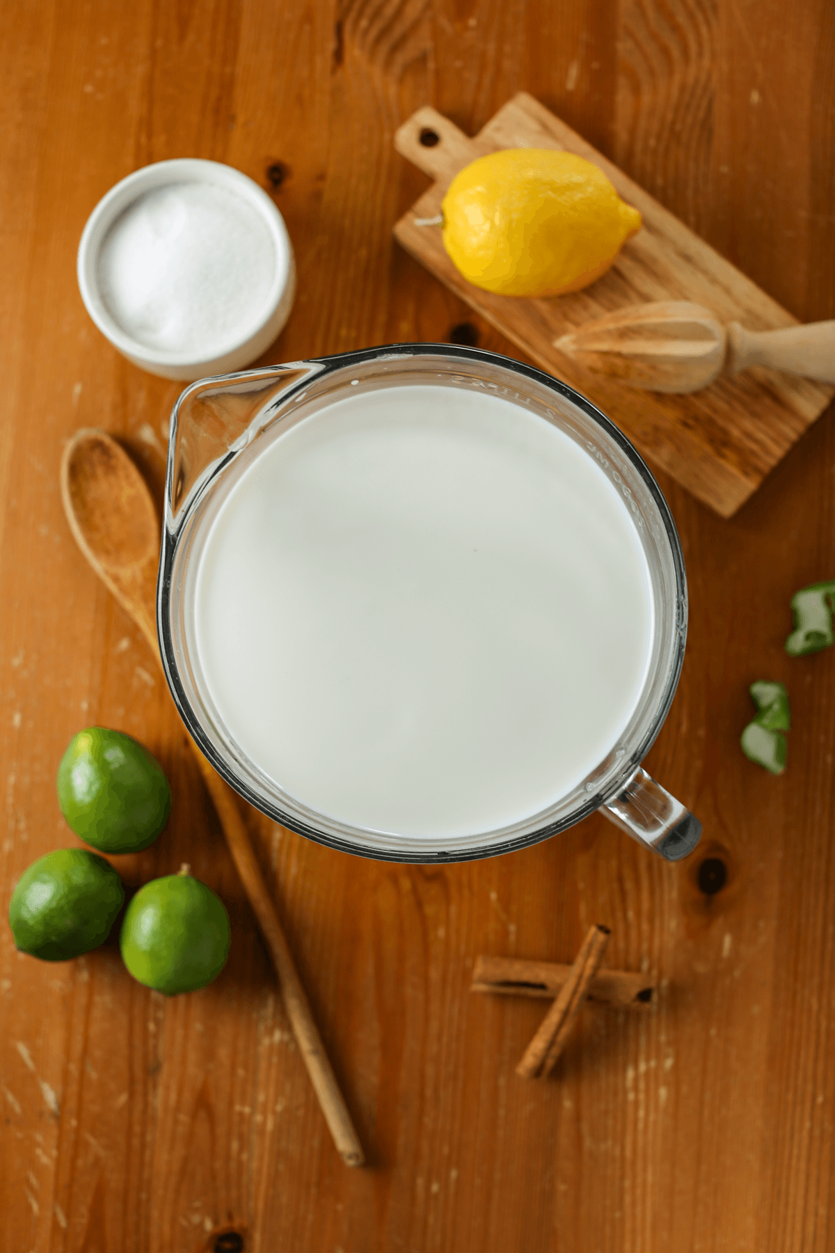 An overlay image where the ingredients needed to make dulce de leche cortada are on a table. There's milk, lemon, sugar, limes, and cinnamon sticks.