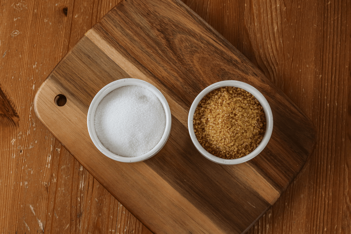 Brown table with two small white bowls. One has white sugar, the other has turbinado sugar.