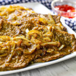 Close up image of browned tender steak with soft julienned onions on top. They're on a white plate.