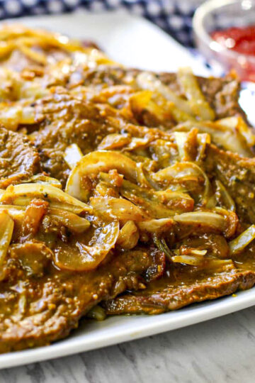 Close up image of browned tender steak with soft julienned onions on top. They're on a white plate.