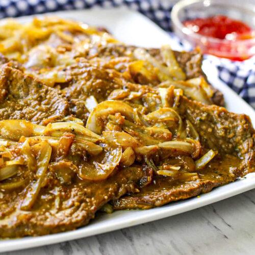 Close up image of browned tender steak with soft julienned onions on top. They're on a white plate.