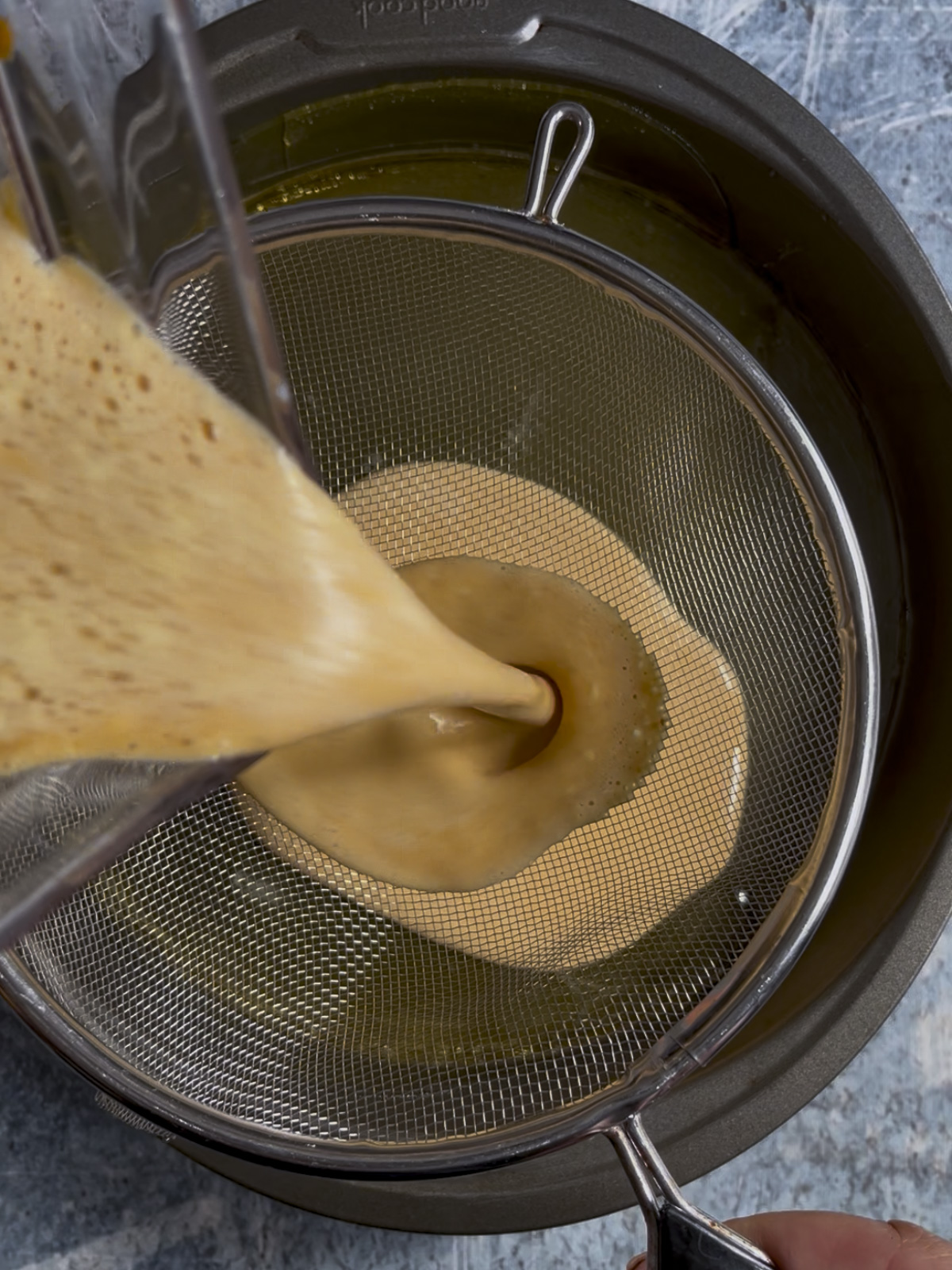 Flan custard is being strained through a sieve into a caramel layered baking mold.